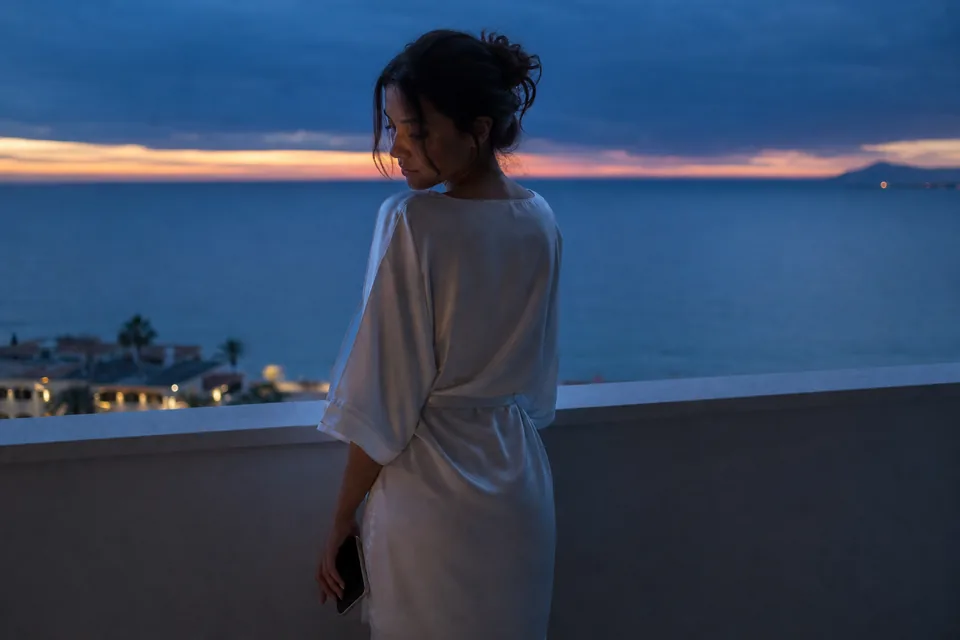 A woman on a balcony above the sea at dusk, face half in shadow, phone in hand